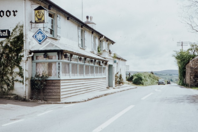 A countryside motel is situated along a quiet rural road. The building features traditional architecture with white walls and a wooden porch. Nearby, there are signs for the AA and RAC, indicating the presence of a hotel. The road is lined with greenery, and there are a few vintage cars parked in the distance.