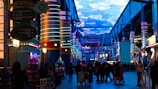 A vibrant indoor complex with illuminated columns and a large digital ceiling display showing a sky pattern. The area is bustling with people, possibly visiting an amusement park or an entertainment venue. There are colorful lights and signage directing to different attractions.