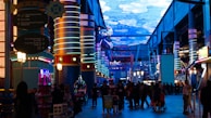 A vibrant indoor complex with illuminated columns and a large digital ceiling display showing a sky pattern. The area is bustling with people, possibly visiting an amusement park or an entertainment venue. There are colorful lights and signage directing to different attractions.
