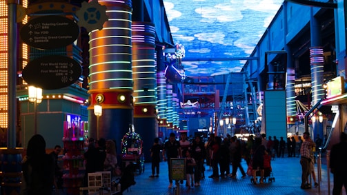 A vibrant indoor complex with illuminated columns and a large digital ceiling display showing a sky pattern. The area is bustling with people, possibly visiting an amusement park or an entertainment venue. There are colorful lights and signage directing to different attractions.