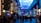 A vibrant indoor complex with illuminated columns and a large digital ceiling display showing a sky pattern. The area is bustling with people, possibly visiting an amusement park or an entertainment venue. There are colorful lights and signage directing to different attractions.