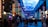 A vibrant indoor complex with illuminated columns and a large digital ceiling display showing a sky pattern. The area is bustling with people, possibly visiting an amusement park or an entertainment venue. There are colorful lights and signage directing to different attractions.