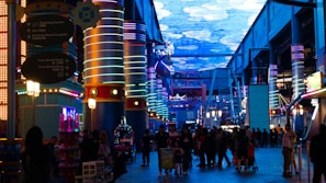 A vibrant indoor complex with illuminated columns and a large digital ceiling display showing a sky pattern. The area is bustling with people, possibly visiting an amusement park or an entertainment venue. There are colorful lights and signage directing to different attractions.