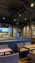 A modern cafe interior featuring wooden tables and gray upholstered chairs arranged neatly. The walls exhibit a combination of vertical wooden slats and framed pictures. A digital menu is displayed behind the counter showcasing various offerings. The ceiling is industrial with exposed ductwork and track lighting providing a warm ambiance.