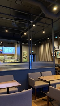 A modern cafe interior featuring wooden tables and gray upholstered chairs arranged neatly. The walls exhibit a combination of vertical wooden slats and framed pictures. A digital menu is displayed behind the counter showcasing various offerings. The ceiling is industrial with exposed ductwork and track lighting providing a warm ambiance.
