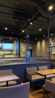 A modern cafe interior featuring wooden tables and gray upholstered chairs arranged neatly. The walls exhibit a combination of vertical wooden slats and framed pictures. A digital menu is displayed behind the counter showcasing various offerings. The ceiling is industrial with exposed ductwork and track lighting providing a warm ambiance.