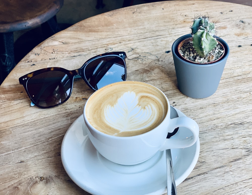 white ceramic cup with saucer beside black sunglasses on brown wooden table