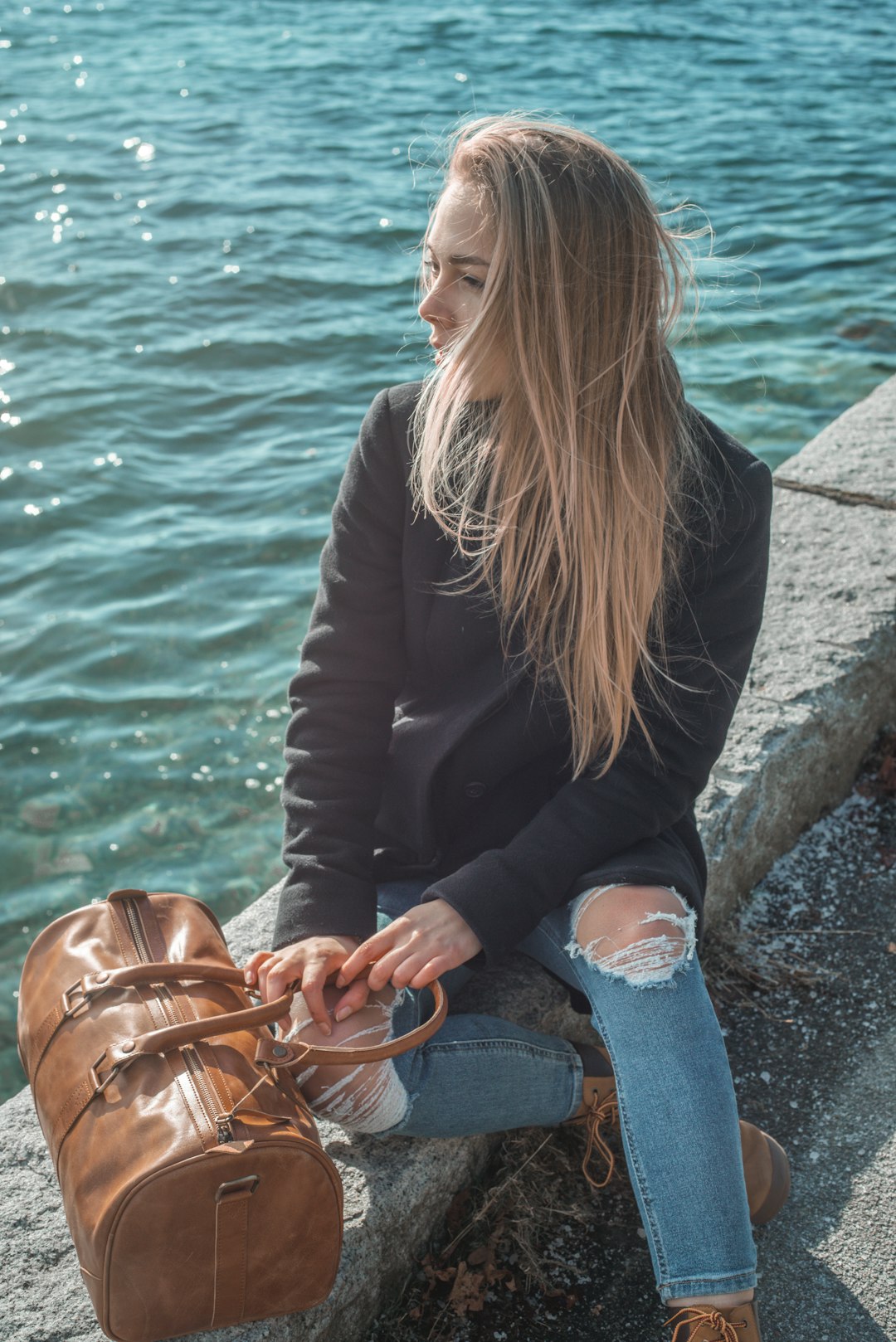 woman in black jacket and blue denim jeans sitting on concrete bench near body of water