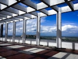 A pergola structure made of white columns and beams stands on a tiled floor, casting geometric shadows. In the background, an expansive landscape with a clear blue sky and puffy clouds stretches out, providing a distant view of greenery and rocky hills.