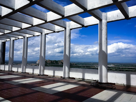 A pergola structure made of white columns and beams stands on a tiled floor, casting geometric shadows. In the background, an expansive landscape with a clear blue sky and puffy clouds stretches out, providing a distant view of greenery and rocky hills.