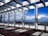 A pergola structure made of white columns and beams stands on a tiled floor, casting geometric shadows. In the background, an expansive landscape with a clear blue sky and puffy clouds stretches out, providing a distant view of greenery and rocky hills.