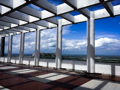 A pergola structure made of white columns and beams stands on a tiled floor, casting geometric shadows. In the background, an expansive landscape with a clear blue sky and puffy clouds stretches out, providing a distant view of greenery and rocky hills.