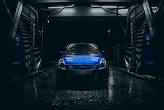 A blue car with raindrops is parked inside an illuminated car wash bay during nighttime. The scene is dark with light reflecting off the wet surfaces, creating a dramatic and moody atmosphere. The car wash bay has visible signage and equipment surrounding the vehicle.
