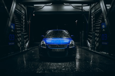 A blue car with raindrops is parked inside an illuminated car wash bay during nighttime. The scene is dark with light reflecting off the wet surfaces, creating a dramatic and moody atmosphere. The car wash bay has visible signage and equipment surrounding the vehicle.