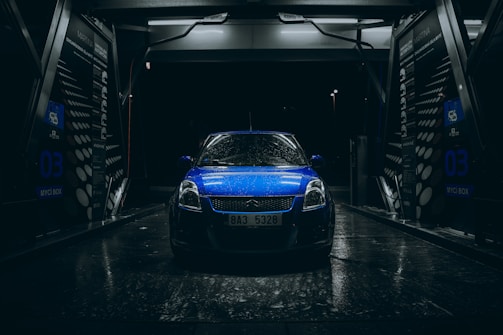 A blue car with raindrops is parked inside an illuminated car wash bay during nighttime. The scene is dark with light reflecting off the wet surfaces, creating a dramatic and moody atmosphere. The car wash bay has visible signage and equipment surrounding the vehicle.