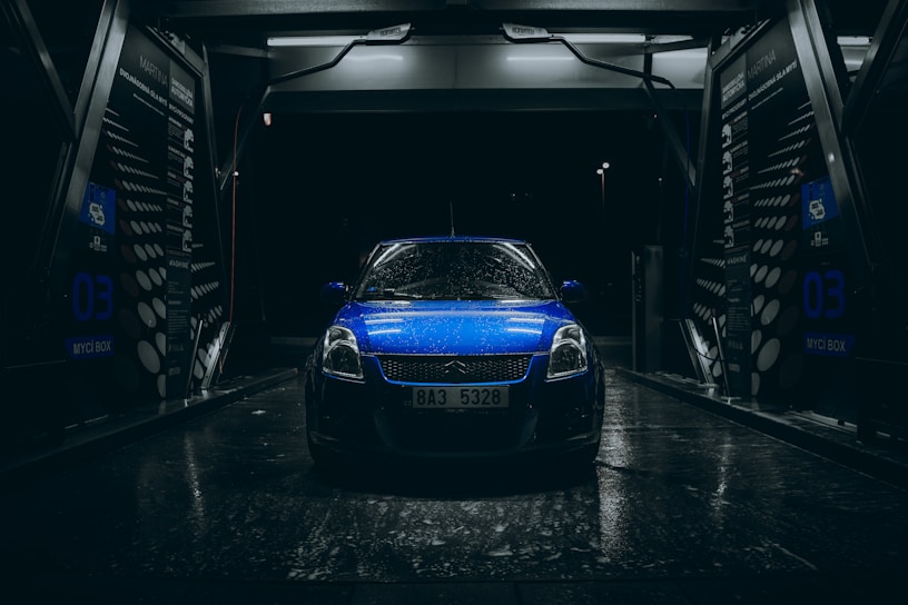 A blue car with raindrops is parked inside an illuminated car wash bay during nighttime. The scene is dark with light reflecting off the wet surfaces, creating a dramatic and moody atmosphere. The car wash bay has visible signage and equipment surrounding the vehicle.