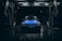 A blue car with raindrops is parked inside an illuminated car wash bay during nighttime. The scene is dark with light reflecting off the wet surfaces, creating a dramatic and moody atmosphere. The car wash bay has visible signage and equipment surrounding the vehicle.
