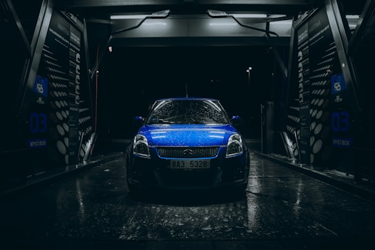 A blue car with raindrops is parked inside an illuminated car wash bay during nighttime. The scene is dark with light reflecting off the wet surfaces, creating a dramatic and moody atmosphere. The car wash bay has visible signage and equipment surrounding the vehicle.