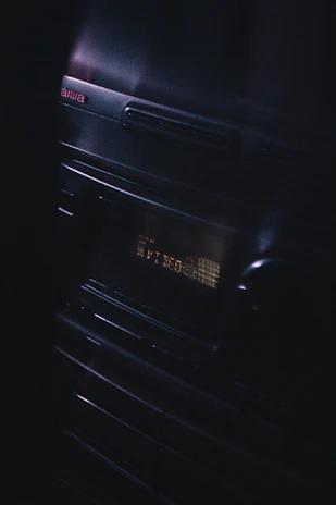 A vintage audio stereo system with a display screen showing text, illuminated in a dimly lit environment. The brand name Aiwa is visible at the top left corner, and the equipment has a sleek, reflective surface with buttons and a knob.