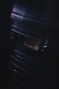 A vintage audio stereo system with a display screen showing text, illuminated in a dimly lit environment. The brand name Aiwa is visible at the top left corner, and the equipment has a sleek, reflective surface with buttons and a knob.