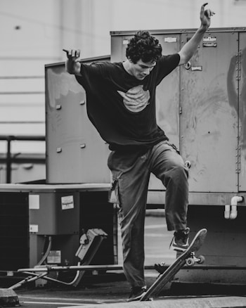A person performing a trick on a skateboard in an urban environment. The individual is wearing a loose-fitting t-shirt and pants, balancing with one foot on the skateboard and the other raised. The background includes industrial elements like metal surfaces and equipment.