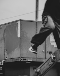 A dynamic black-and-white photograph capturing a person performing a trick with a skateboard in front of industrial metal units. The image focuses on motion, with the skateboard diagonally positioned, highlighting energy and movement.