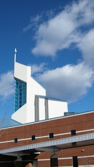 A modern church building featuring a tall, angular white section with a cross atop it, and a brick section with rectangular windows. The clear blue sky with scattered, fluffy clouds forms the backdrop.
