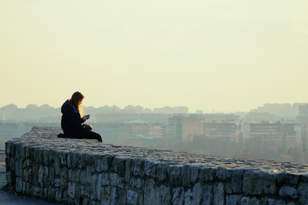 woman in black jacket sitting on gray concrete wall during daytime