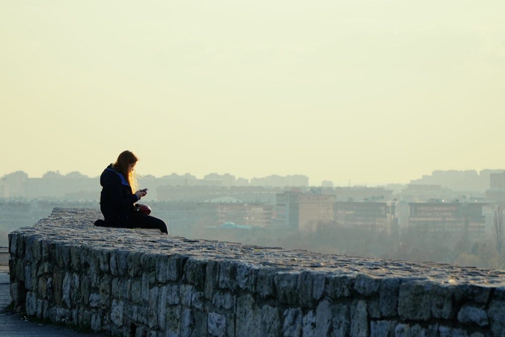 Femme en veste noire assise sur un mur de béton gris pendant la journée