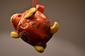 A decorative plush object shaped like a pig, covered in a red fabric with intricate golden patterns. The plush has yellow feet, ears, and snout, with a loop at the top for hanging.