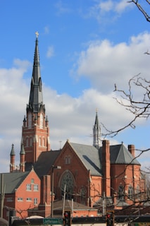 A large, red-brick church with a tall spire topped by a cross stretches into a blue sky with scattered clouds. The building features intricate architectural details, including multiple steeples and arched windows. The foreground shows bare tree branches and the top of a street sign that reads 'Jefferson'.