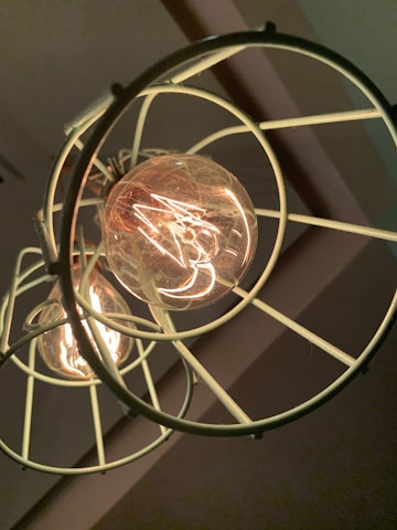 A close-up view of an incandescent light bulb enclosed within a circular metal cage suspended from the ceiling. The bulb's filaments are glowing warmly, casting a soft light and creating intricate patterns of light and shadow on the surrounding surfaces.