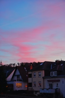 A residential scene with a row of houses featuring pitched roofs and visible chimneys. The sky above is painted with a stunning gradient of blue transitioning into pink and purple hues, indicating sunset or sunrise. Trees are silhouetted against the colorful sky in the background, and warm lights glow from some of the windows, adding a cozy atmosphere.