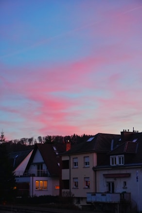 A residential scene with a row of houses featuring pitched roofs and visible chimneys. The sky above is painted with a stunning gradient of blue transitioning into pink and purple hues, indicating sunset or sunrise. Trees are silhouetted against the colorful sky in the background, and warm lights glow from some of the windows, adding a cozy atmosphere.