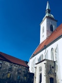 Tall white church with a green spire and clock tower set against a clear blue sky. The building features arched windows and a detailed entrance. Adjacent to the church is an old wall with murals.