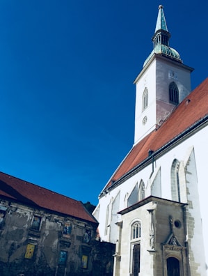 Tall white church with a green spire and clock tower set against a clear blue sky. The building features arched windows and a detailed entrance. Adjacent to the church is an old wall with murals.