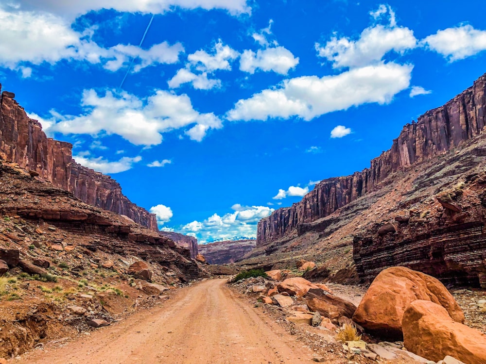 brown dirt road between rocky mountains under blue sky and white clouds during daytime