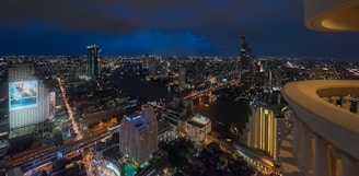 A vibrant cityscape at night with numerous high-rise buildings illuminated, a river running through the center, and glowing billboards adding color to the scene. The sky is dark with a tint of blue, and streets are bustling with lights, indicating a lively urban atmosphere.