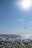 A rocky shore overlooks the sea, with waves gently crashing onto the stones. A kiteboarder is visible sailing across the water, accompanied by a small sailboat in the distance. The sun shines brightly, casting a shimmering light over the ocean surface and illuminating the clear blue sky.