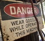 A weathered safety sign is affixed to a wooden surface, with the word 'DANGER' in bold white letters against a red oval background. Below, the text advises wearing goggles while operating a machine. The sign shows signs of aging, such as rust and discoloration. A cable or wire runs beside the sign.