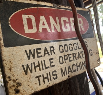 A weathered safety sign is affixed to a wooden surface, with the word 'DANGER' in bold white letters against a red oval background. Below, the text advises wearing goggles while operating a machine. The sign shows signs of aging, such as rust and discoloration. A cable or wire runs beside the sign.