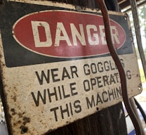 A weathered safety sign is affixed to a wooden surface, with the word 'DANGER' in bold white letters against a red oval background. Below, the text advises wearing goggles while operating a machine. The sign shows signs of aging, such as rust and discoloration. A cable or wire runs beside the sign.
