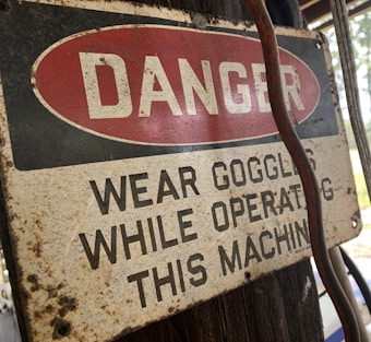 A weathered safety sign is affixed to a wooden surface, with the word 'DANGER' in bold white letters against a red oval background. Below, the text advises wearing goggles while operating a machine. The sign shows signs of aging, such as rust and discoloration. A cable or wire runs beside the sign.
