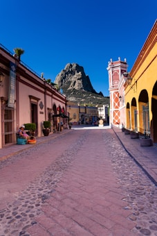 A cobblestone street in a quaint town with colorful buildings on either side. The sky is clear and blue, providing a vibrant backdrop. In the distance, a prominent rocky mountain rises, adding to the scenic beauty. There are potted plants along the street and baskets of goods near a building entrance. A few signs in Spanish are visible, hinting at a possibly Latin American location.