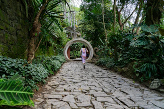 girl in pink jacket walking on pathway between green trees during daytime