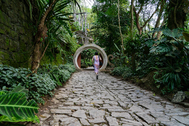 girl in pink jacket walking on pathway between green trees during daytime