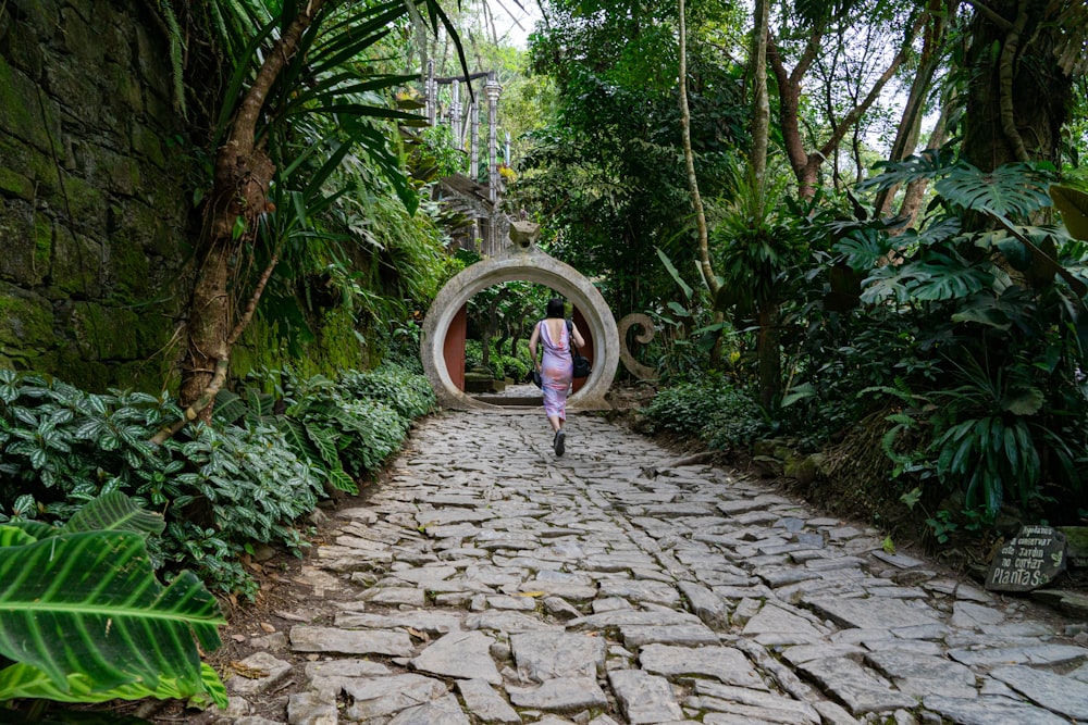 girl in pink jacket walking on pathway between green trees during daytime