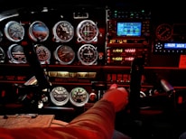 A cockpit panel filled with various dials, gauges, and digital displays, indicating flight information. The lighting is a mix of bright LED displays and red ambient light, highlighting the details of the controls and instruments. A person's hand is gripping the yoke, suggesting active control of the aircraft.