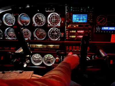 A cockpit panel filled with various dials, gauges, and digital displays, indicating flight information. The lighting is a mix of bright LED displays and red ambient light, highlighting the details of the controls and instruments. A person's hand is gripping the yoke, suggesting active control of the aircraft.