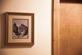 A framed illustration of two squirrels is hung on a beige wall. The artwork is bordered by a wooden frame, showing the squirrels in a natural pose on a pale background. The scene is captured next to a wooden door, which occupies part of the right side of the image.